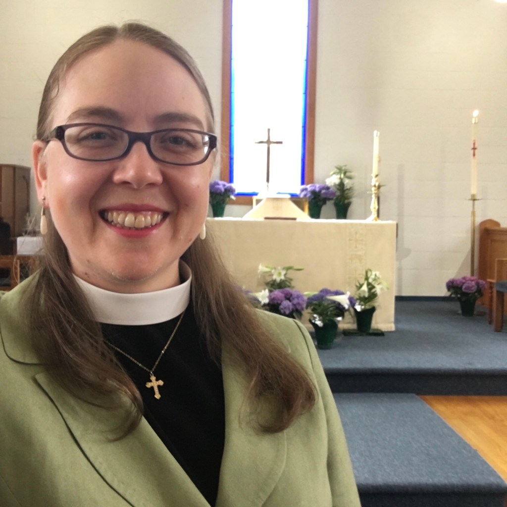 Diana wearing a clerical collar and green blazer in front of an altar decorated with Easter flowers 